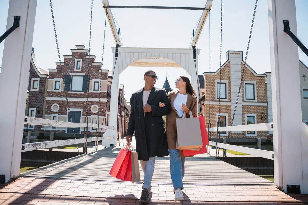 pareja interracial con estilo sonriendo el uno al otro y caminando con bolsas de compras a través del puente - Foto, imagen
