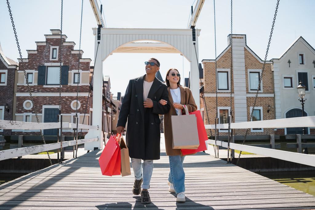 longitud completa de sonriente pareja interracial en gafas de sol y abrigos caminando con bolsas de compras en el puente - Foto, imagen