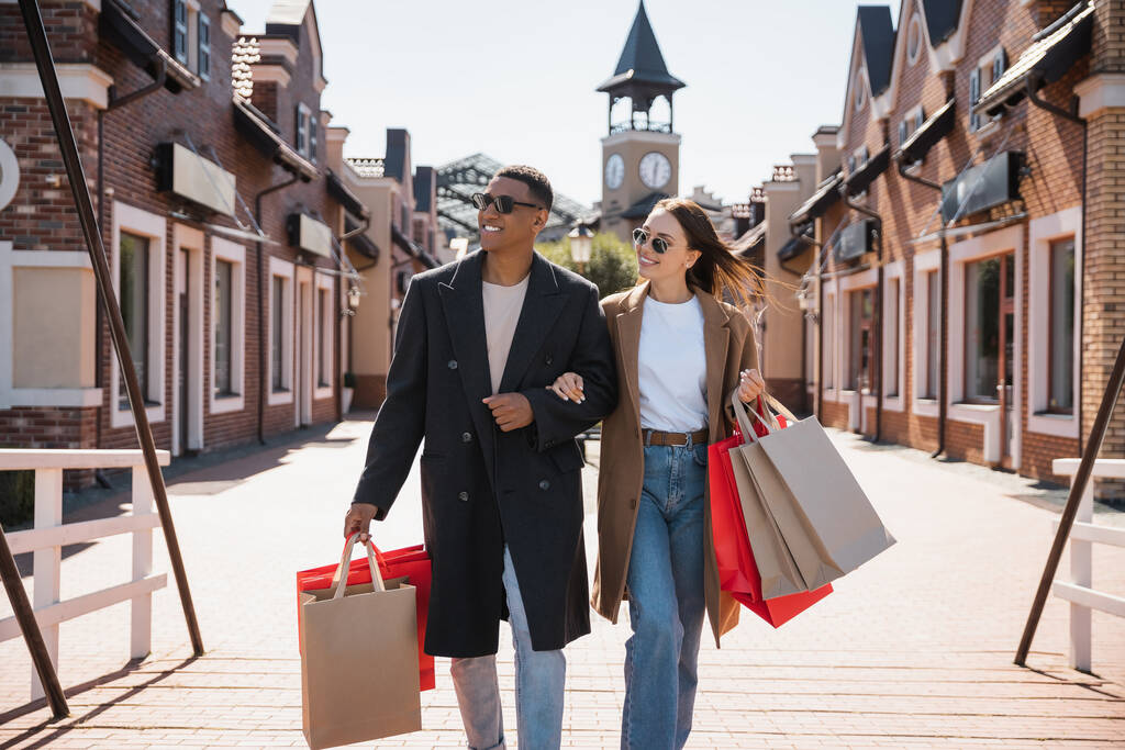 pareja interracial en abrigos de moda y gafas de sol caminando con bolsas de compras y mirando hacia el puente - Foto, Imagen