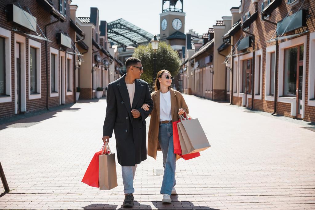 pełna długość wieloetnicznej pary w płaszczach i okularach przeciwsłonecznych spacerujących z torbami na zakupy na ulicy miejskiej - Zdjęcie, obraz
