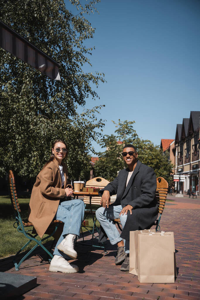 happy multiethnic couple in coats and sunglasses sitting near coffee to go and shopping bags in street cafe - Photo, Image