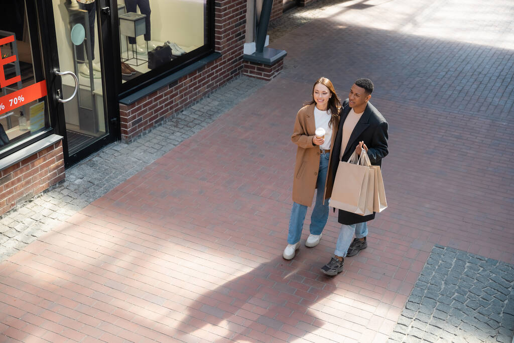 vista de ángulo alto de la elegante pareja multicultural con bolsas de compras y taza de papel caminando junto a la tienda con vitrinas - Foto, Imagen