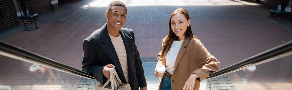 trendiges multiethnisches Paar mit Einkaufstaschen und Kaffee zum Anlächeln in die Kamera auf Rolltreppe, Banner - Foto, Bild