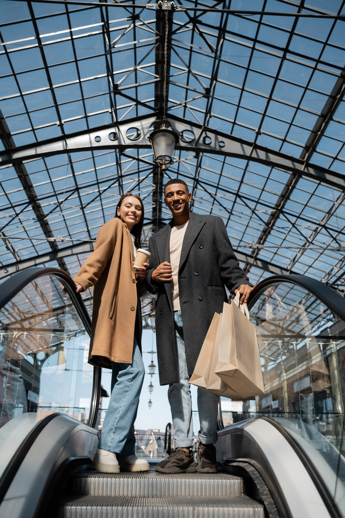 trendy woman with paper cup and african american man with shopping bags smiling at camera on escalator under glass roof - Photo, Image