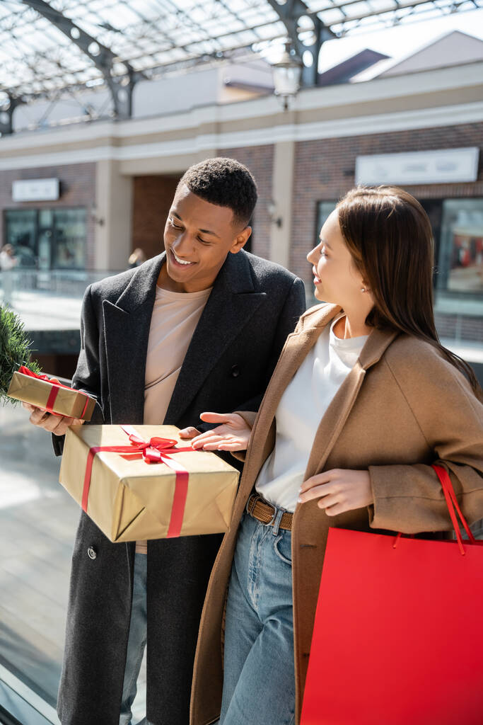 陽気なアフリカ系アメリカ人男性ショッピングバッグと若いガールフレンドの近くにギフトボックスを保持 - 写真・画像