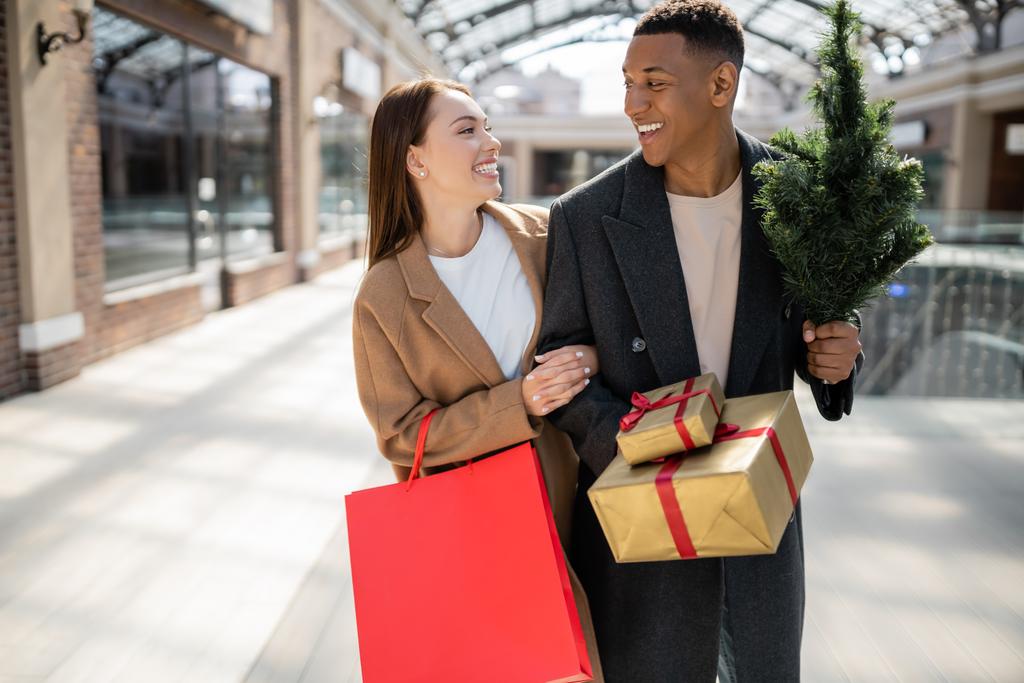 stílusos interracial pár kabátban tartó újévi ajándékok és kis karácsonyfa, miközben mosolyognak egymásra - Fotó, kép