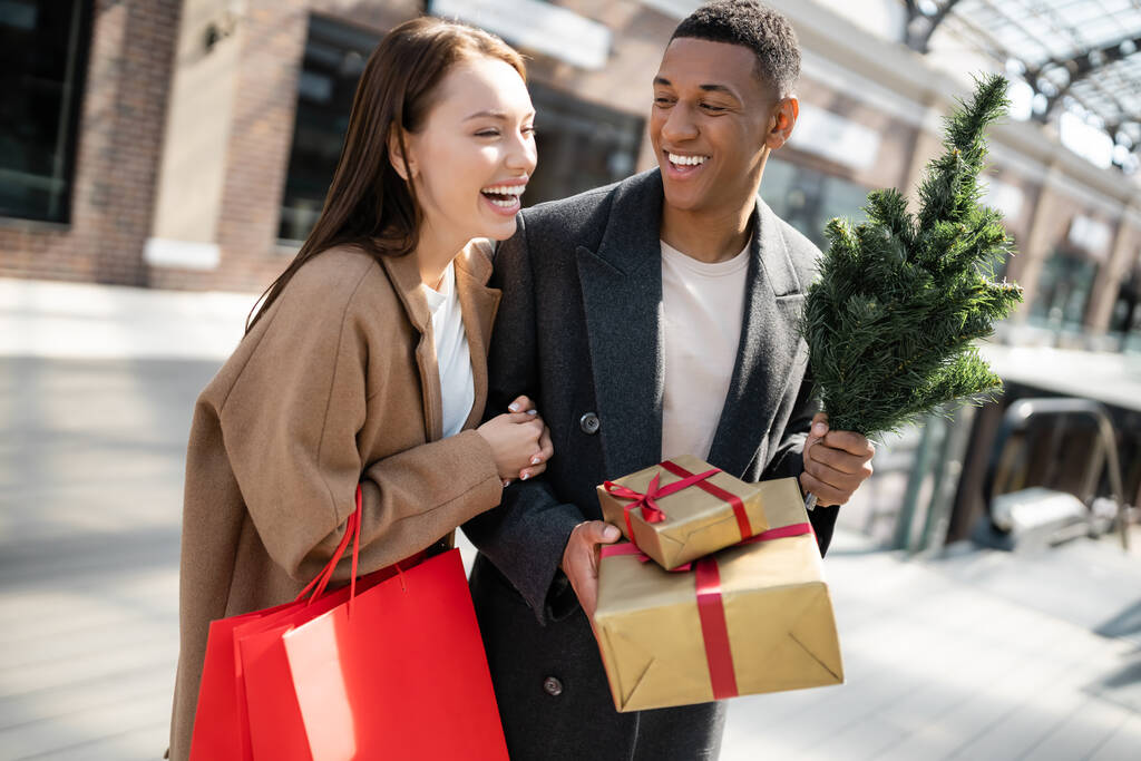 femme excitée avec des sacs à provisions riant près élégant homme afro-américain tenant des cadeaux et petit arbre de Noël - Photo, image