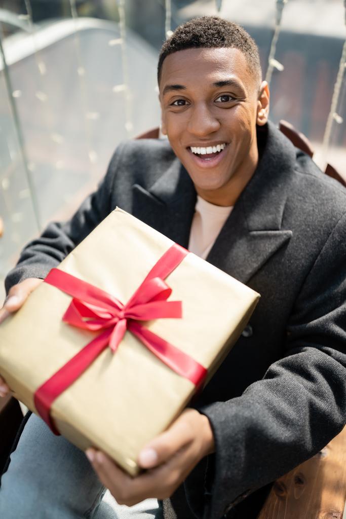 hombre afroamericano emocionado con caja de regalo mirando a la cámara al aire libre - Foto, Imagen
