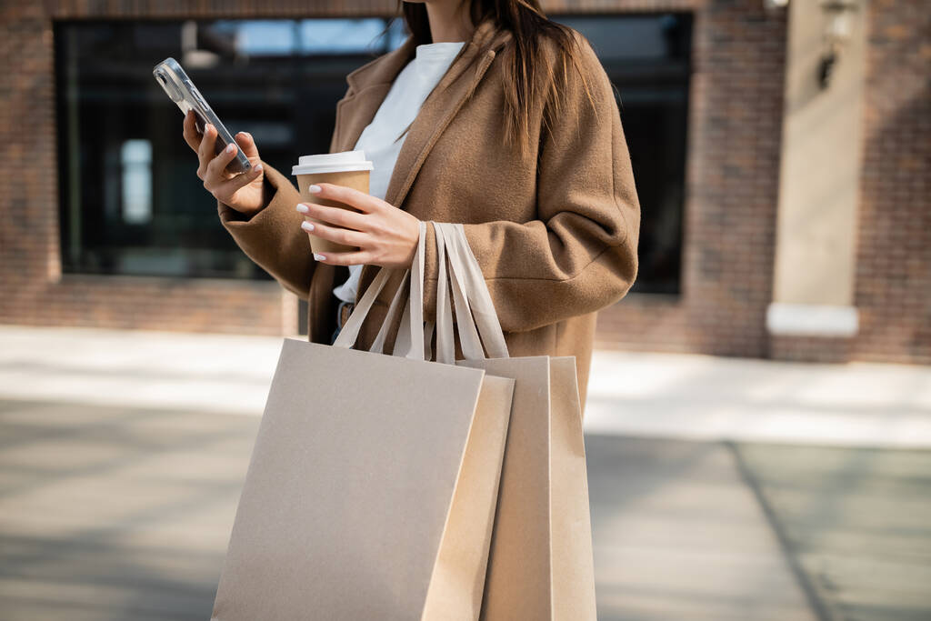 スマートフォンで女性を切り取り街中で紙コップ付きのショッピングバッグを持ち - 写真・画像