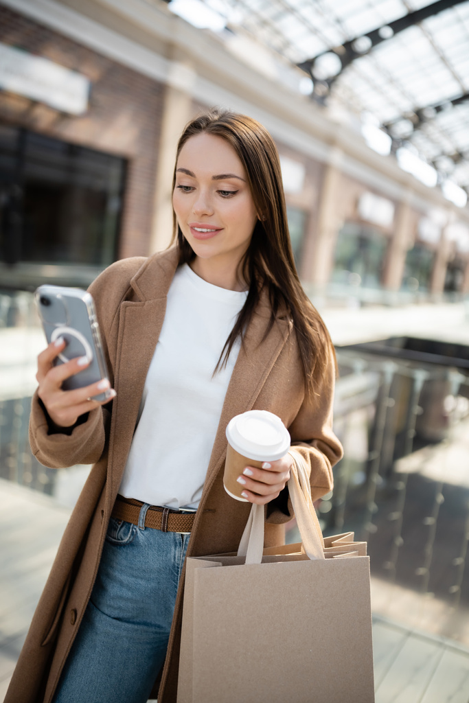 mulher morena na moda segurando copo de papel e sacos de compras enquanto olha para o smartphone - Foto, Imagem
