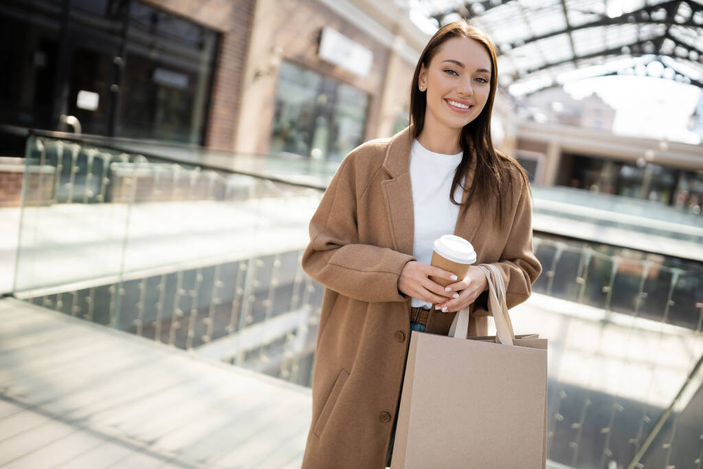boldog és elegáns nő elvitelre ital és bevásárló táskák nézi kamera közelében elmosódott bevásárlóközpont - Fotó, kép