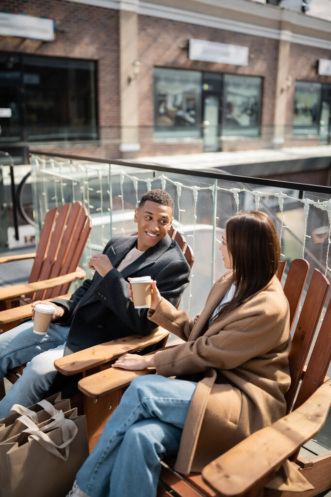 весёлая многонациональная пара в пальто, сидящая с кофе, чтобы подойти к сумкам и размытому торговому центру - Фото, изображение