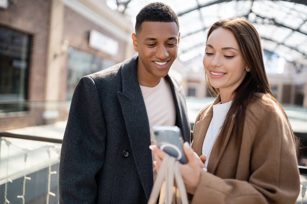 jong brunette vrouw tonen mobiele telefoon naar glimlachen Afrikaans amerikaans vriendje op wazig straat - Foto, afbeelding