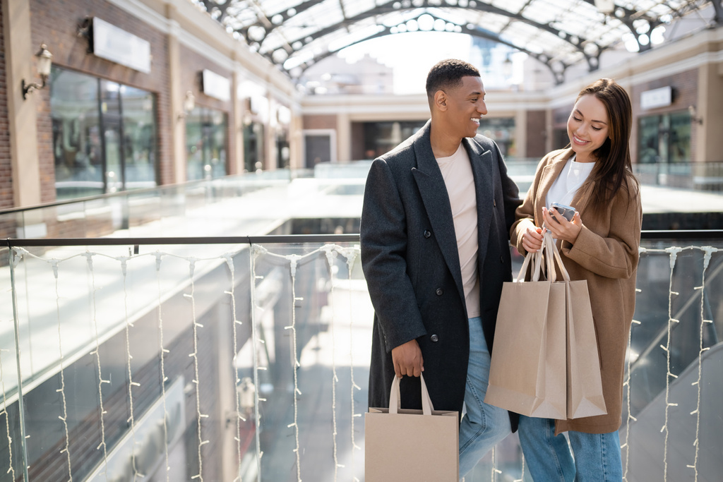 mujer feliz mirando el teléfono inteligente cerca de novio afroamericano con bolsas de compras  - Foto, imagen
