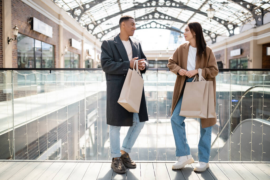 полная длина межрасовой пары в пальто и джинсах, держащих сумки с покупками и смотрящих друг на друга возле торгового центра - Фото, изображение