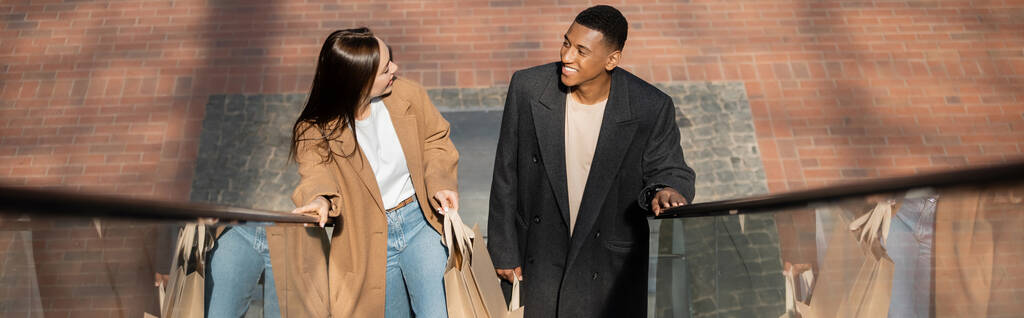 vista ad alto angolo di coppia multietnica sorridente con shopping bag che si guardano su scala mobile, banner - Foto, immagini