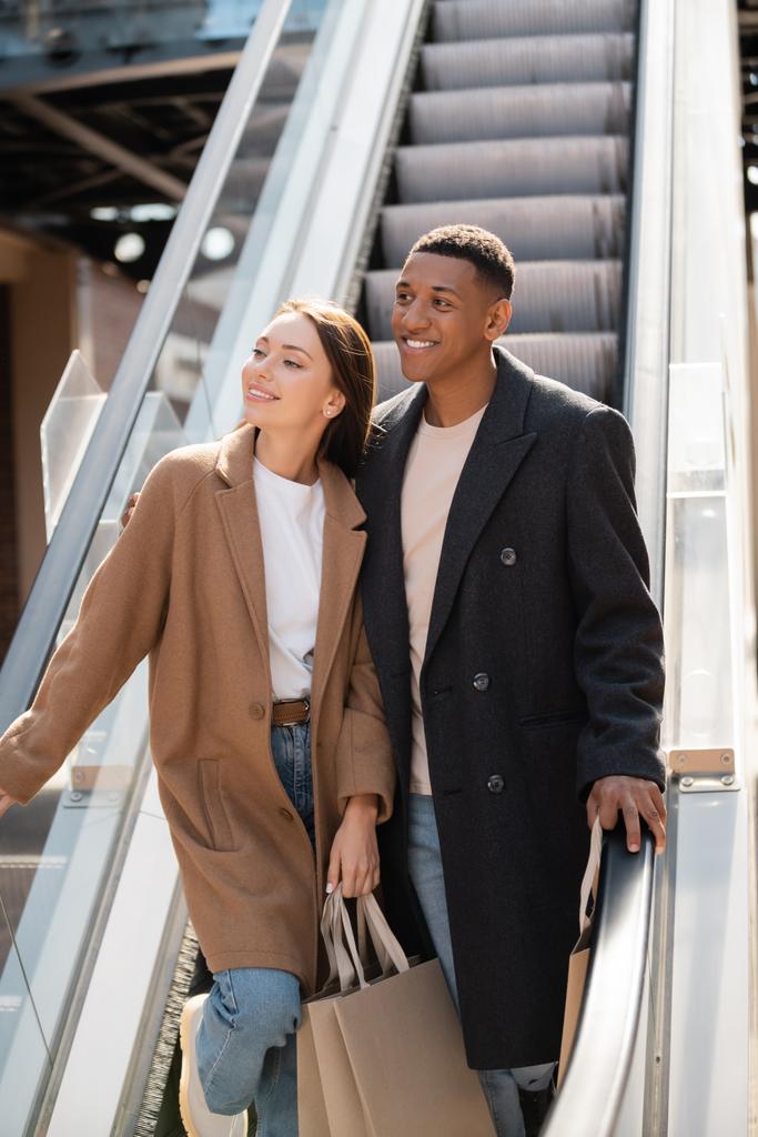 šťastný a stylový multietnický pár s nákupními taškami odvrací pohled na eskalátoru - Fotografie, Obrázek