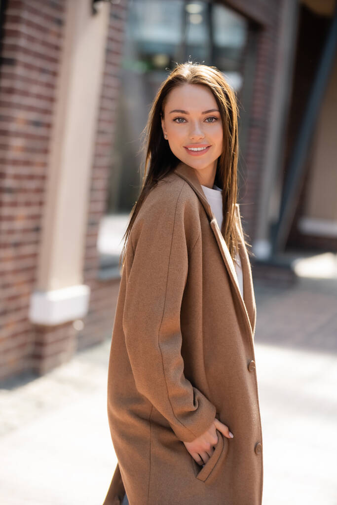 mladá a hezká brunetka žena stojící s rukou v kapse béžového kabátu a usmívající se na kameru - Fotografie, Obrázek