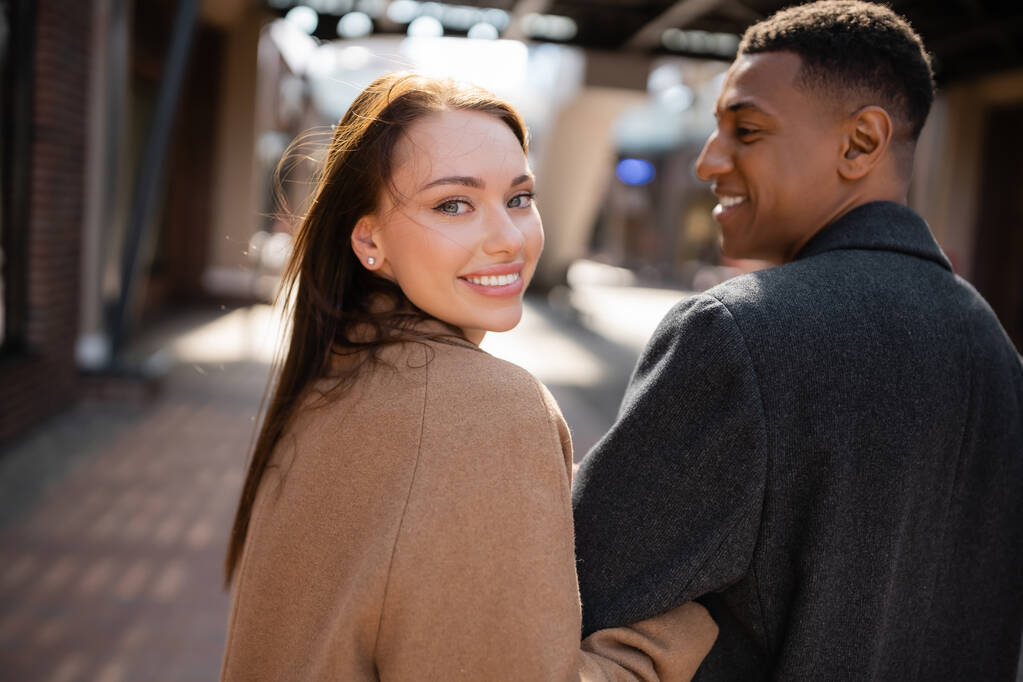 pretty and happy woman looking at camera near african american boyfriend on blurred street - Photo, Image