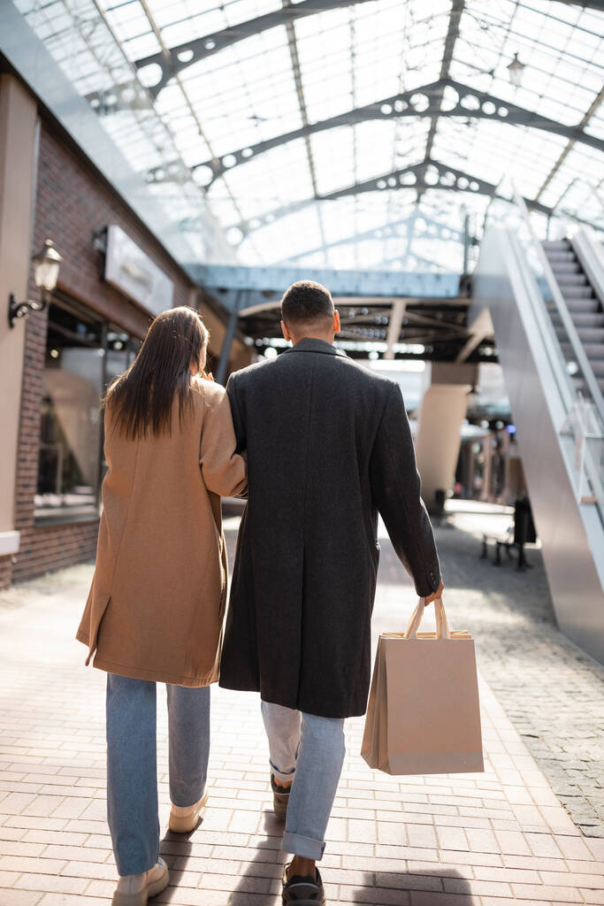Rückansicht eines afrikanisch-amerikanischen Mannes mit Einkaufstüten, der mit seiner brünetten Freundin auf der städtischen Straße spaziert - Foto, Bild
