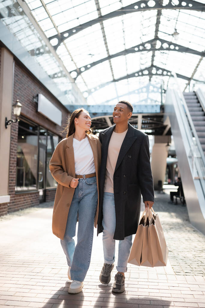 piena lunghezza di donna elegante sorridente al fidanzato afro-americano con borse della spesa vicino al centro commerciale - Foto, immagini