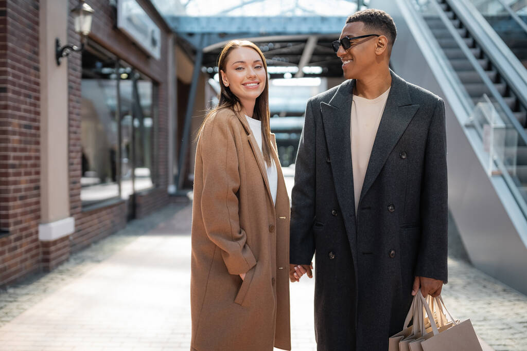 mujer complacida con la mano en el bolsillo del abrigo cogido de la mano con el hombre afroamericano con estilo con bolsas de compras - Foto, Imagen