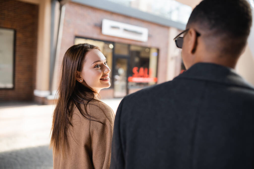 happy brunette woman looking at african american boyfriend on blurred foreground in city - Photo, Image