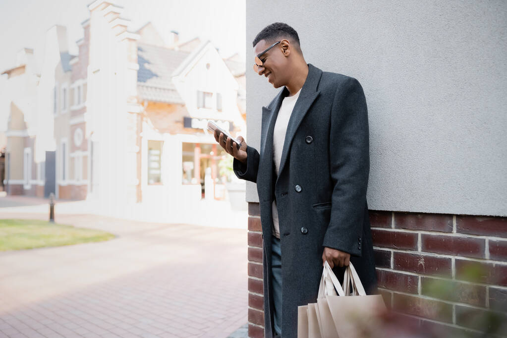 веселий афроамериканський чоловік в пальто і сонцезахисних окулярах тримає сумки і спілкується на мобільному телефоні біля будівлі
 - Фото, зображення