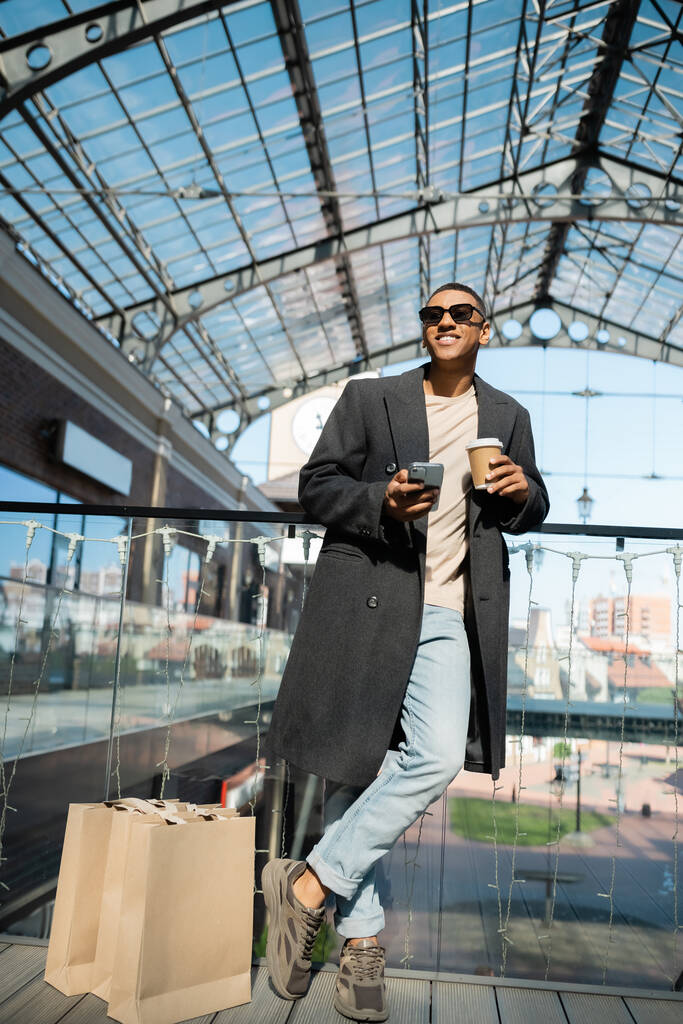 Kahveli ve akıllı telefonlu Afro-Amerikan bir adam alışveriş çantaları ve alışveriş merkezlerinin yanında duruyor. - Fotoğraf, Görsel