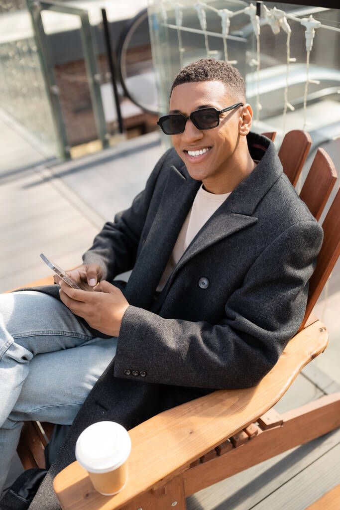 african american man in coat and sunglasses sitting with mobile phone and smiling at camera on urban street - Photo, Image