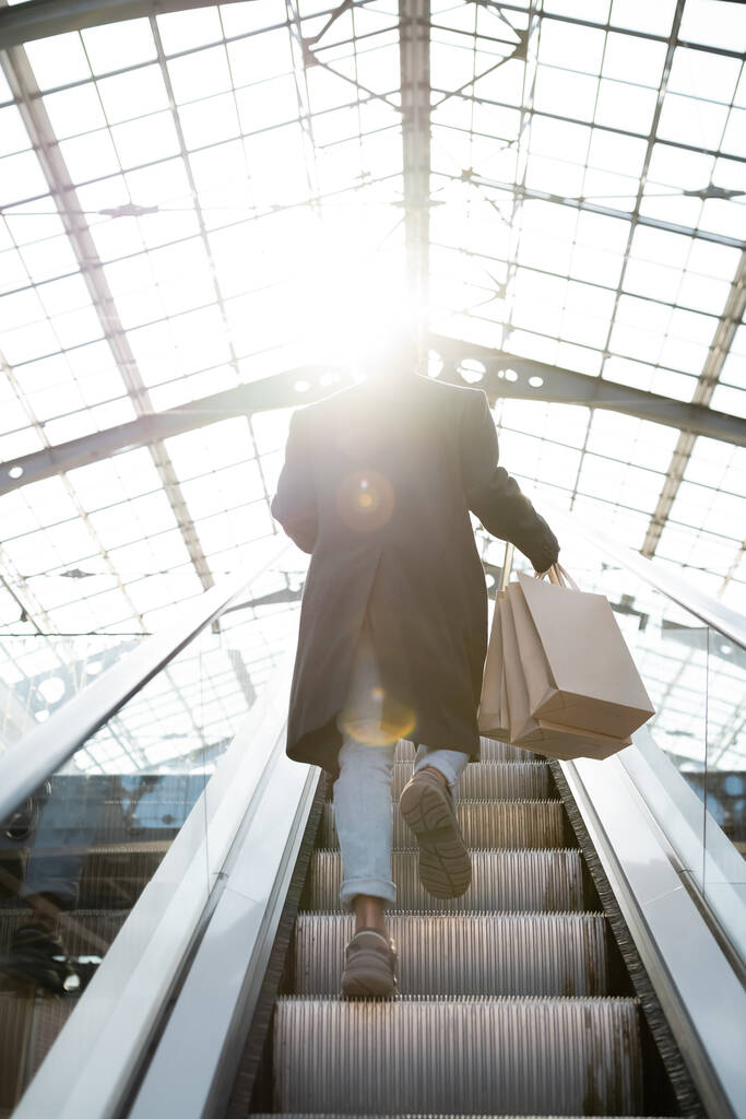 visão traseira do homem americano africano com sacos de compras andando na escada rolante em sol - Foto, Imagem