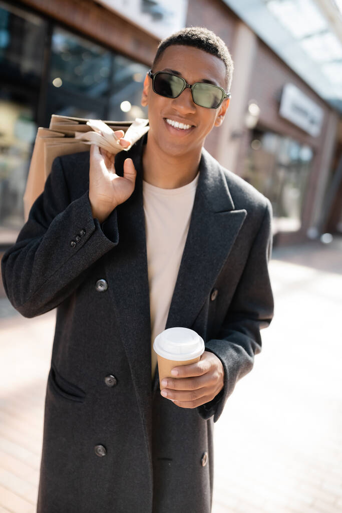 trendiger afrikanisch-amerikanischer Mann in Sonnenbrille mit Getränken zum Mitnehmen und Einkaufstüten, während er auf der Stadtstraße in die Kamera lächelt - Foto, Bild