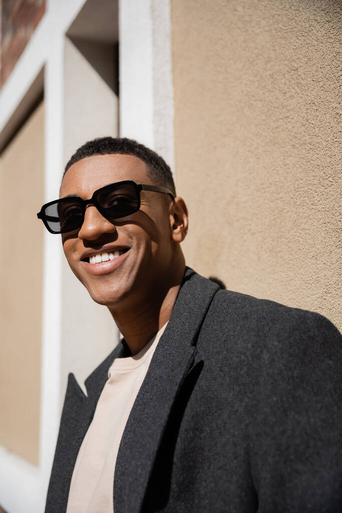 radostný afroameričan v kabátě a slunečních brýlích stojící u zdi a dívající se do kamery - Fotografie, Obrázek