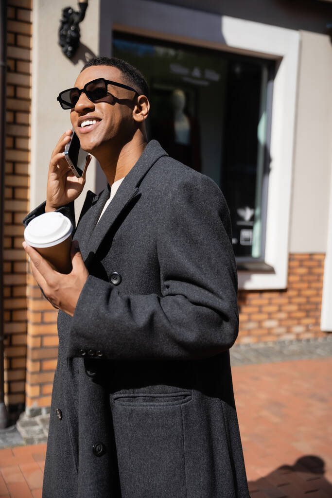 χαρούμενος Αφροαμερικάνος με παλτό και γυαλιά ηλίου κρατώντας καφέ για να πάει και να μιλήσει στο smartphone στο δρόμο - Φωτογραφία, εικόνα