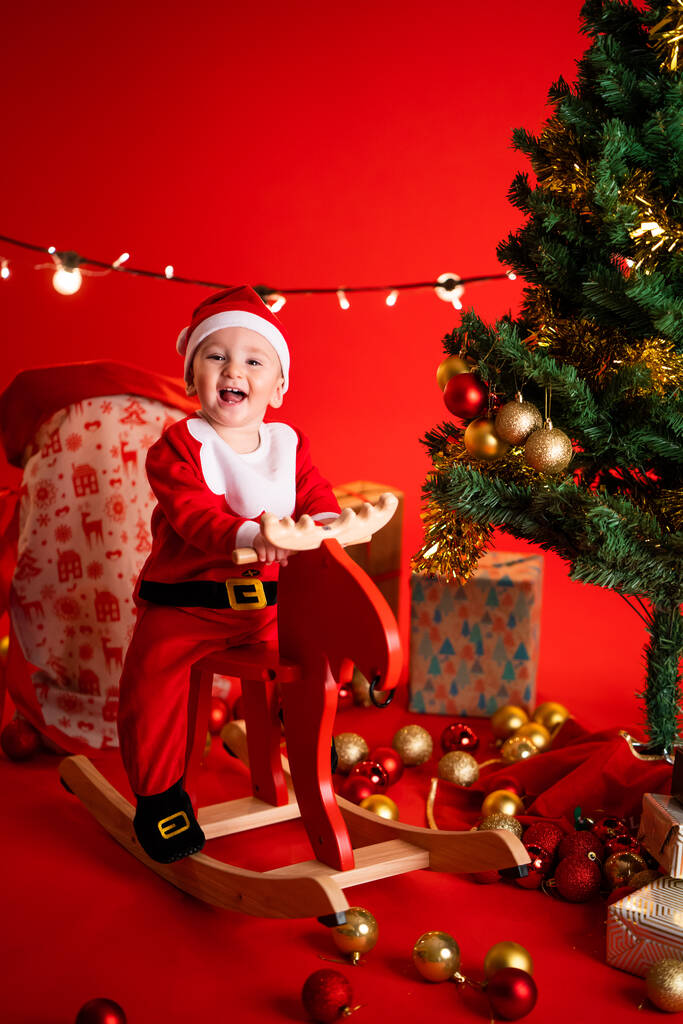Full length of adorable little child in Santa hat and costume sitting on rocking toy and looking at camera in studio with Christmas tree and decorations on red background - Photo, Image