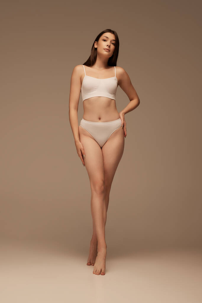 Full-length portret van jonge slanke vrouw poseren in wit ondergoed geïsoleerd over donker beige studio achtergrond. Concept van schoonheid, lichaams- en huidverzorging, gezondheid, plastische chirurgie, cosmetica, sport - Foto, afbeelding