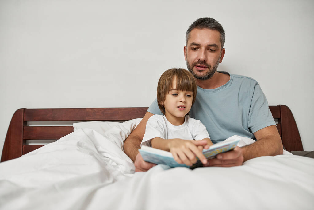 Vader knuffelt en leest boek aan kleine zoon in bed thuis. Jonge blanke familierelatie en samen tijd doorbrengen. Vaderschap en ouderschap. Huishoudelijke vrijetijdsbesteding, hobby en entertainment - Foto, afbeelding
