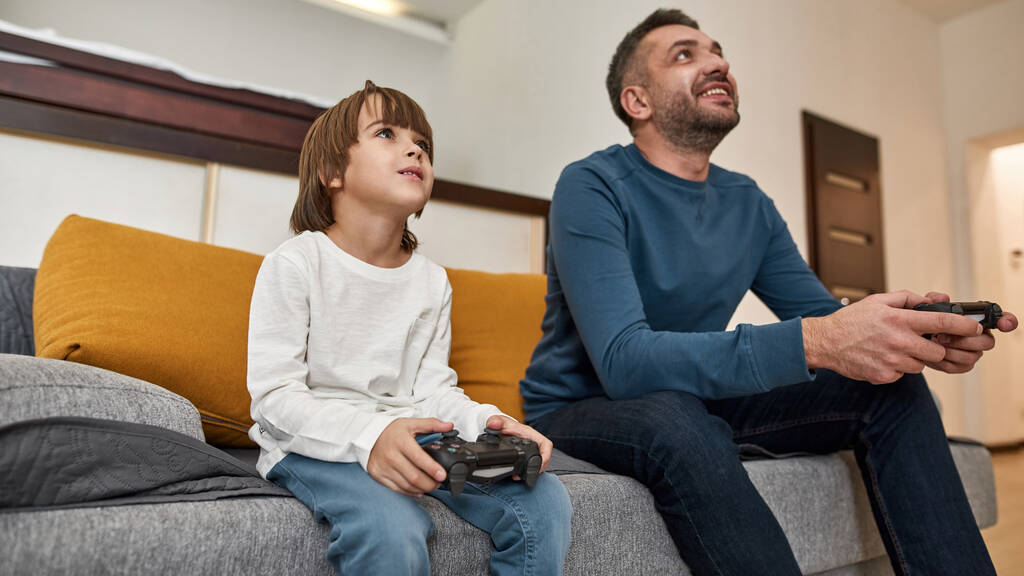 Niño y padre caucásicos jugando videojuegos con joysticks en el sofá en casa. Niño enfocado y hombre sonriente pasando tiempo juntos. Paternidad y paternidad. Ocio doméstico y entretenimiento - Foto, imagen