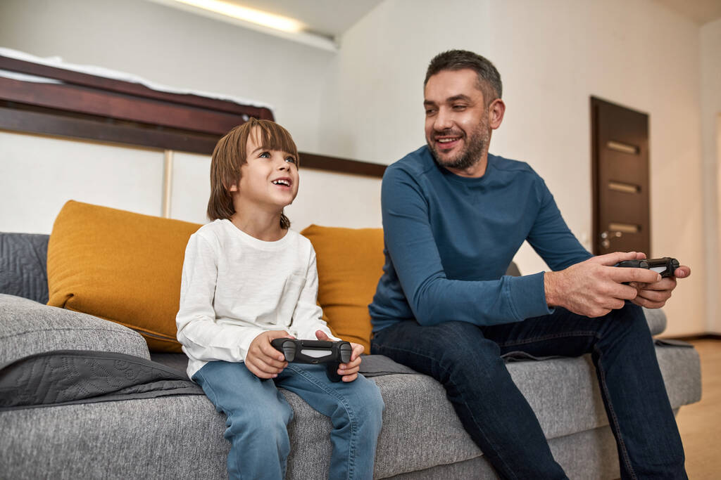 Caucasian little boy and father play video game with joysticks on sofa at home. Smiling man look at concentrated boy. Fatherhood and parenting. Family relationship. Domestic leisure and entertainment - Photo, Image