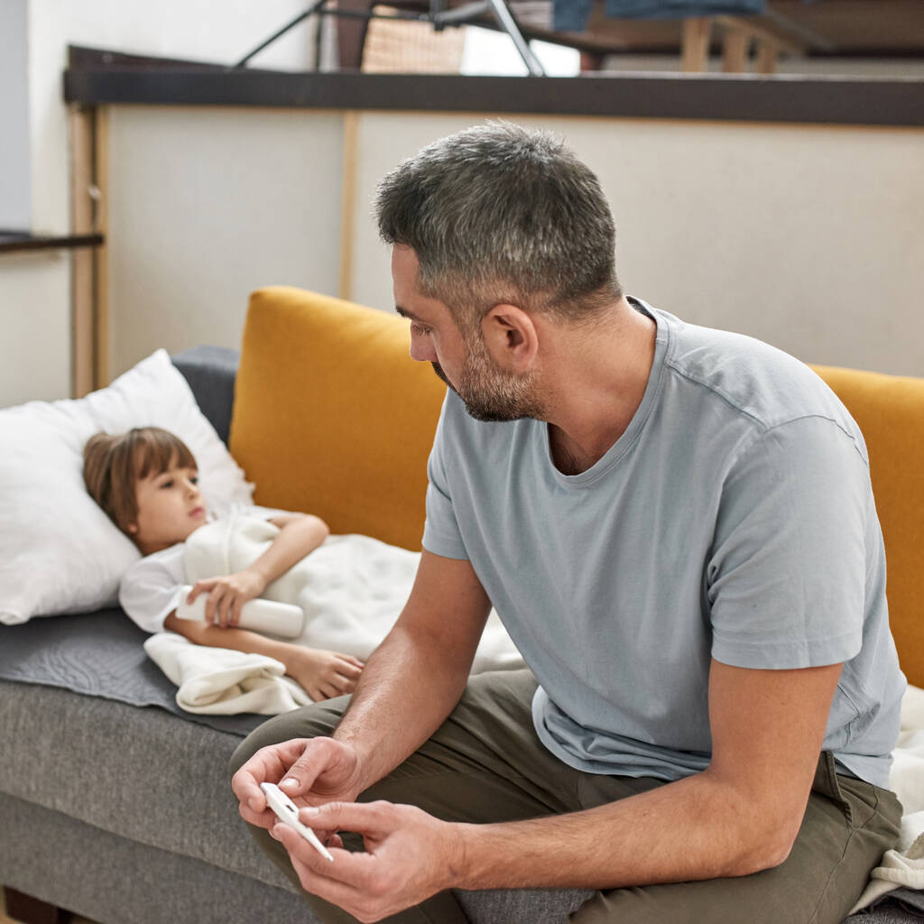 Father holding thermometer and looking at his sick little son with heat lying on sofa at home. Fatherhood and parenting. Domestic lifestyle. Health care. Young caucasian family relationship - Photo, Image