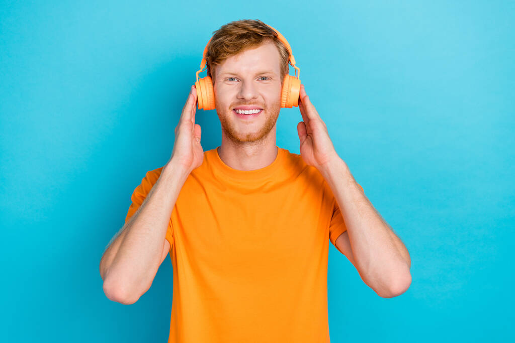 Foto de lindo chico encantador vestido naranja ropa brazos toque auriculares disfrutando de música encantadora aislado om cyan color fondo. - Foto, Imagen