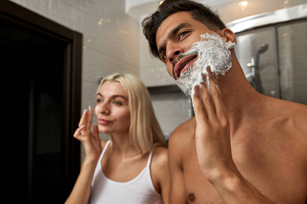 Nahöstlicher Mann schmiert Bart mit Rasierschaum, während kaukasisches Mädchen morgens im Badezimmer das Gesicht mit Kosmetikcreme schmiert. Häuslicher Lebensstil. Gesichtspflege. Junges multiethnisches Paar - Foto, Bild