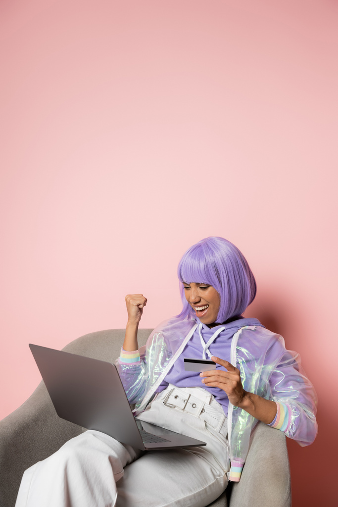 excitée femme afro-américaine en perruque violette en utilisant un ordinateur portable tout en tenant la carte de crédit et faire des achats en ligne le vendredi noir sur rose  - Photo, image