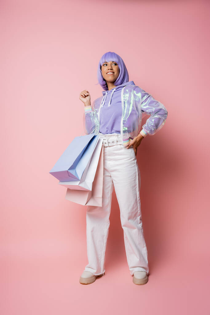幸せなアフリカ系アメリカ人女性の完全な長さ透明ジャケットを保持ショッピングバッグとピンクの上にヒップで手で立って  - 写真・画像