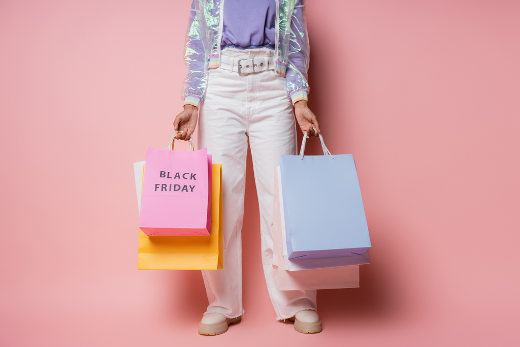 cropped άποψη της γυναίκας σε διαφανές σακάκι και λευκό παντελόνι κρατώντας τσάντες για ψώνια με μαύρο γράμματα Παρασκευή σε ροζ  - Φωτογραφία, εικόνα