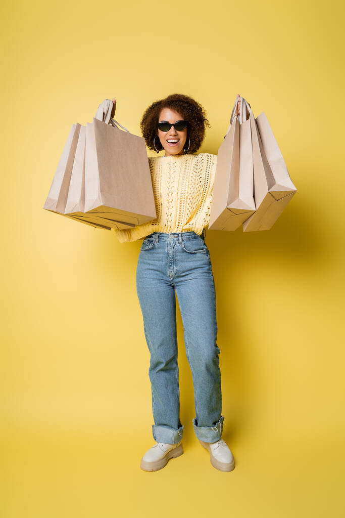 in voller Länge positive afrikanisch-amerikanische Frau mit Sonnenbrille und Einkaufstaschen auf gelb - Foto, Bild