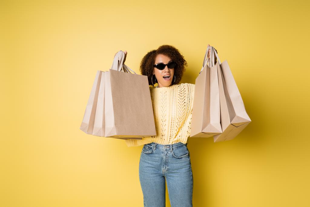 excited african american woman in stylish sunglasses holding shopping bags on yellow - Photo, Image