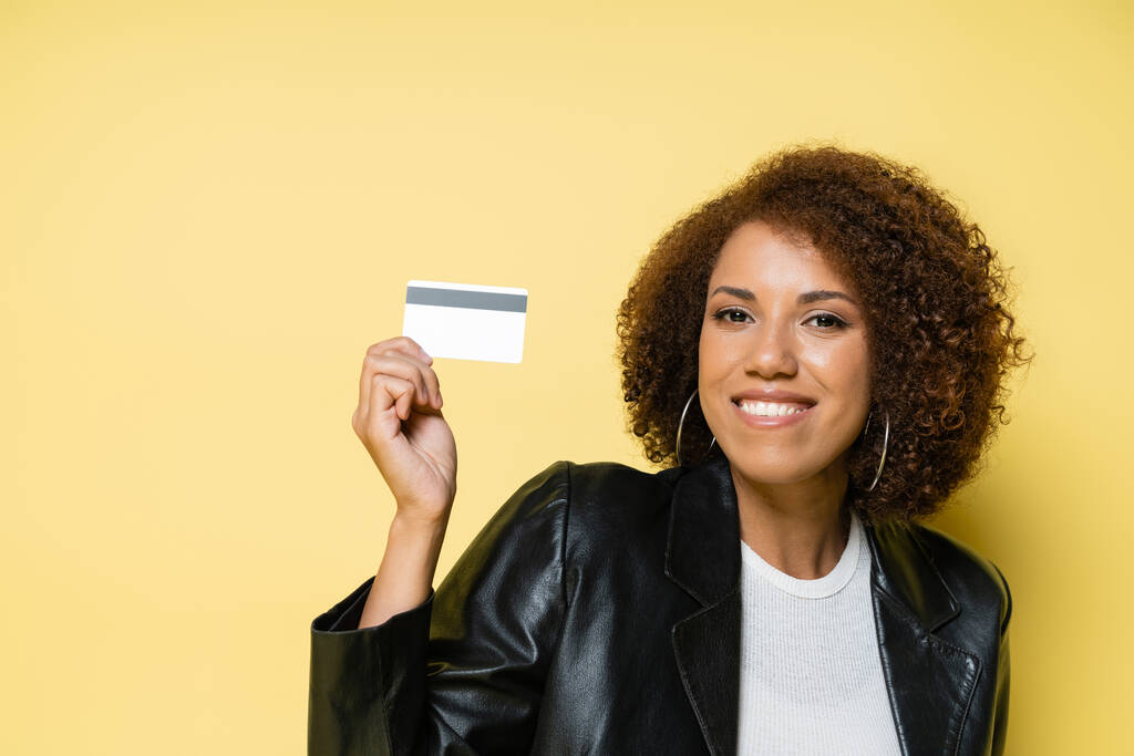 vidám és fiatal afro-amerikai nő bőrdzsekiben kezében hitelkártya elszigetelt sárga  - Fotó, kép