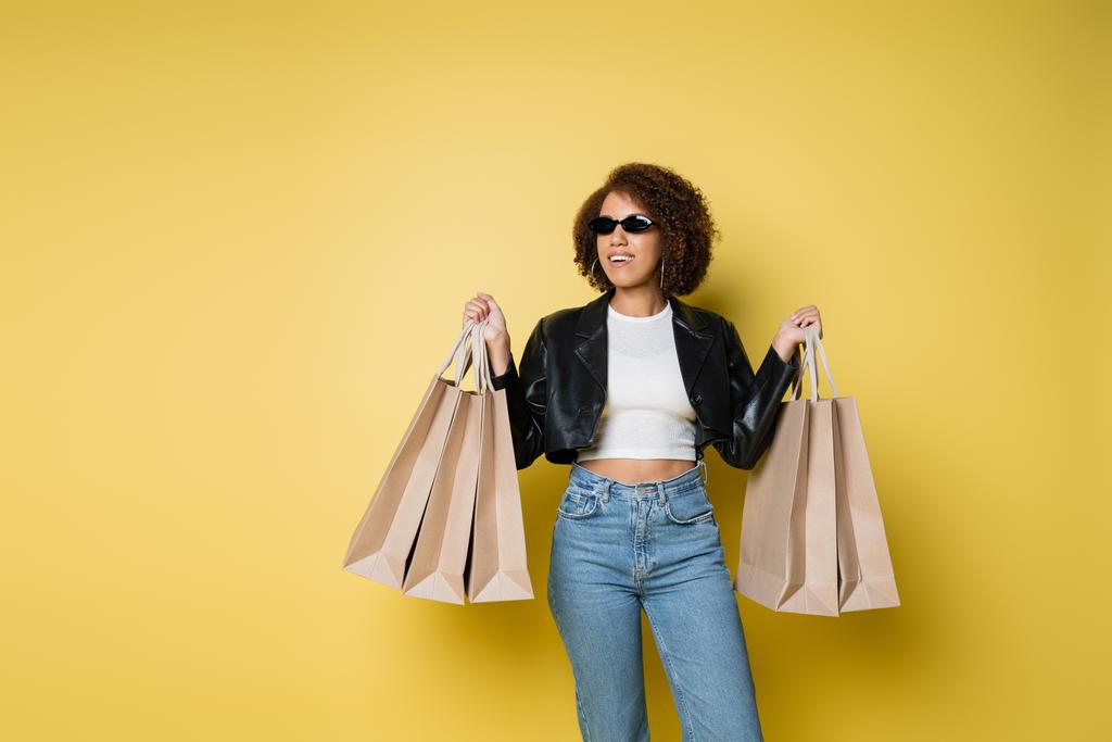Güneş gözlüklü ve deri ceketli neşeli Afro-Amerikalı kadın sarıda siyah cuma günü alışveriş torbaları tutuyordu. - Fotoğraf, Görsel