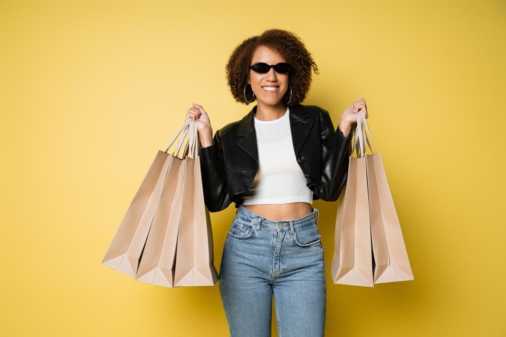 positiivinen afrikkalainen amerikkalainen nainen aurinkolasit ja nahkatakki tilalla ostoskassit keltainen - Valokuva, kuva